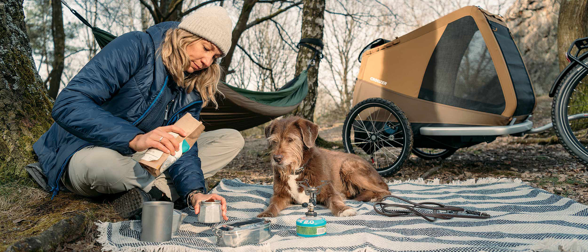 In a forest, a woman makes coffee on a picnic blanket with her dog watching; Croozer Dog trailer attached to bike nearby and a hammock between trees behind.